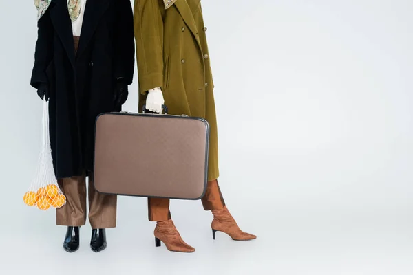 Partial view of women in coats holding vintage suitcase and net bag with oranges on grey background — Stock Photo