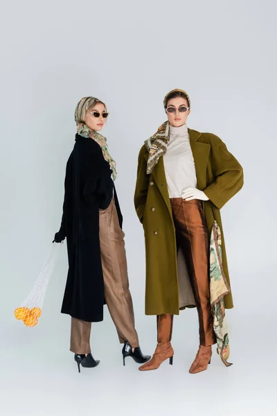 Pleine longueur de femme à la mode avec des oranges dans un sac en filet près d'un ami debout avec la main à la taille sur fond gris — Photo de stock