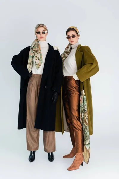 Longitud completa de las mujeres en abrigos de moda y gafas de sol de pie sobre fondo gris - foto de stock