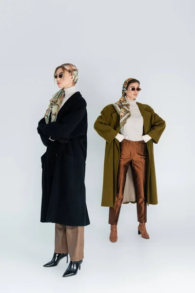 Longitud completa de las mujeres en abrigos y gafas de sol posando con los brazos cruzados y las manos en las caderas sobre fondo gris - foto de stock