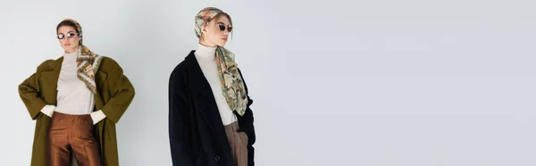 Stylish women in sunglasses and coats posing isolated on grey with copy space, banner — Stock Photo