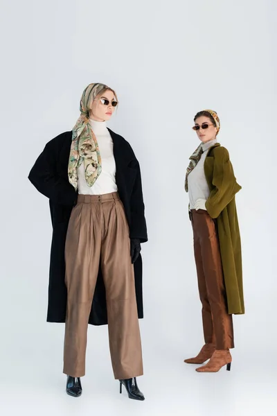 Longitud completa de las mujeres elegantes en abrigos y gafas de sol posando sobre fondo gris - foto de stock