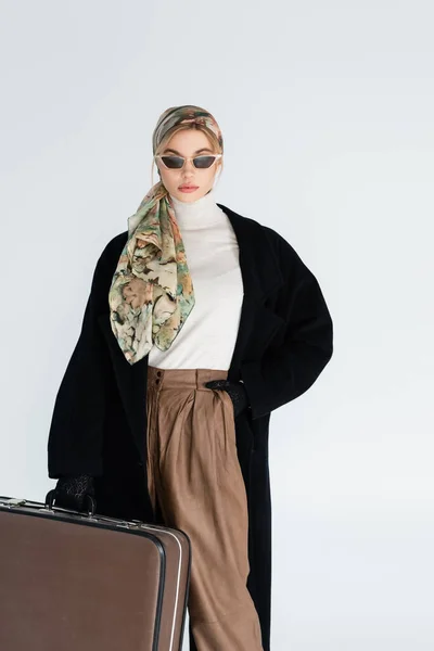 Femme en manteau noir, lunettes de soleil tendance et foulard debout avec valise vintage isolée sur gris — Photo de stock