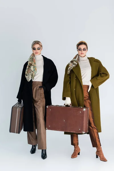 Longitud completa de las mujeres con estilo en abrigos y gafas de sol posando con maletas vintage sobre fondo gris - foto de stock