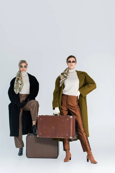 Mujeres con abrigos elegantes y pañuelos elegantes posando con maletas retro sobre fondo gris - foto de stock