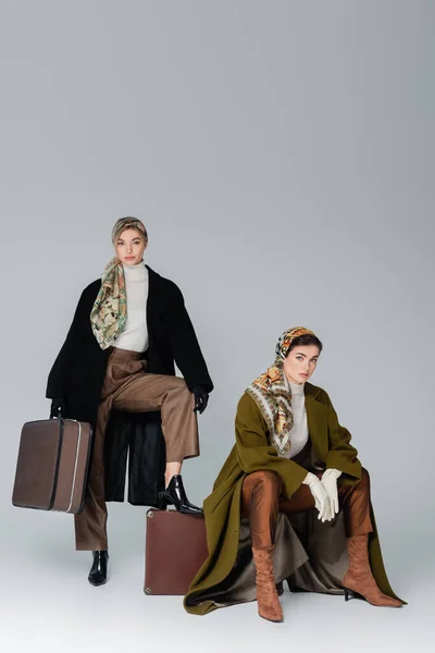 Женщина в черном пальто и платке возле стильного друга сидит на винтажном чемодане на сером фоне — стоковое фото
