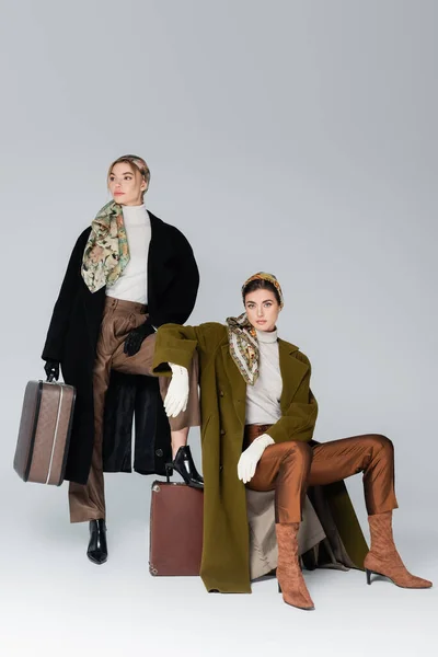 Модная женщина в пальто и перчатках сидит на винтажном чемодане рядом с другом на сером фоне — стоковое фото