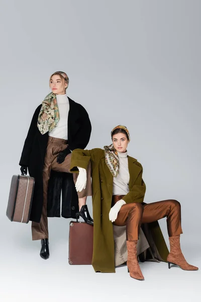 Femme à la mode en manteau et gants assis sur une valise vintage proche ami sur fond gris — Photo de stock