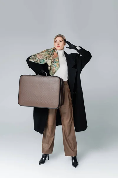 Full length of woman in black coat posing with retro suitcase and hand near head on grey — Stock Photo