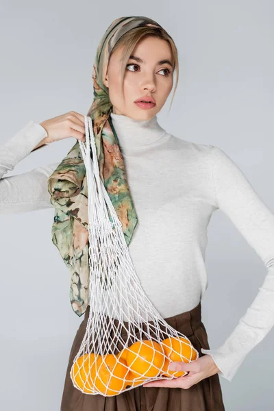 Femme en mouchoir élégant posant avec des oranges fraîches dans un sac en filet isolé sur gris — Photo de stock