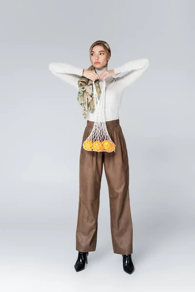 Полная длина женщины в бежевых брюках позирует с апельсинами в сетке мешок на сером — стоковое фото