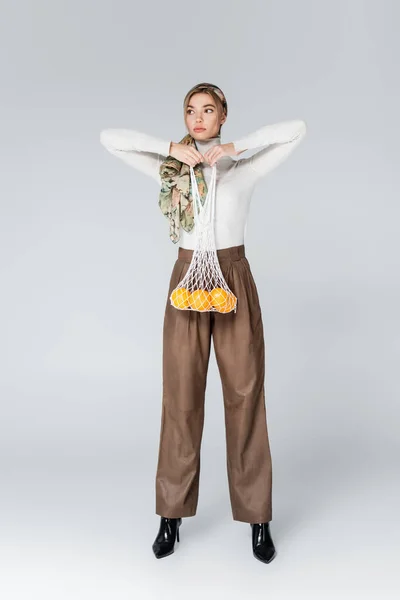 Longueur totale de la femme en pantalon beige posant avec des oranges dans un sac en maille sur gris — Photo de stock