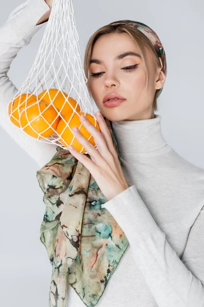 Jolie femme en foulard posant avec des oranges fraîches dans un sac en filet isolé sur gris — Photo de stock