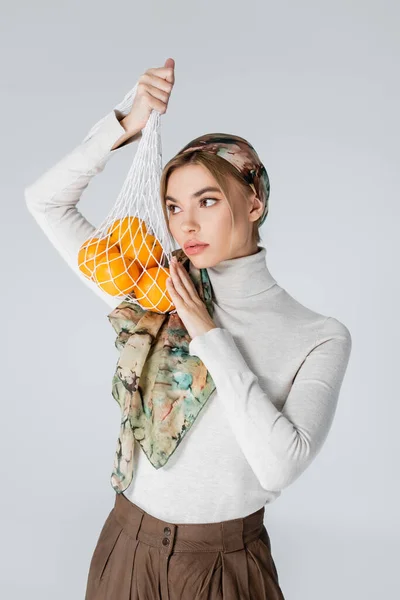 Jolie femme en col roulé blanc et foulard tenant sac filet avec oranges près du visage isolé sur gris — Photo de stock