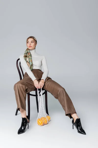 Longitud completa de la mujer en pantalones beige sentado en silla con naranjas maduras en bolsa de red sobre fondo gris - foto de stock