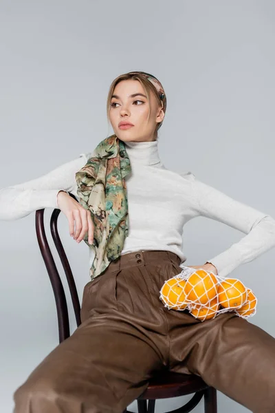 Trendy woman in kerchief sitting on chair with oranges in net bag isolated on grey — Stock Photo