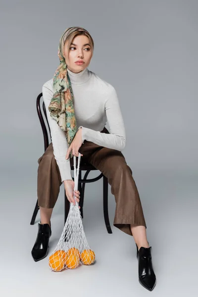 Woman in kerchief and trousers looking away while sitting with fresh oranges in net bag on grey background — Stock Photo