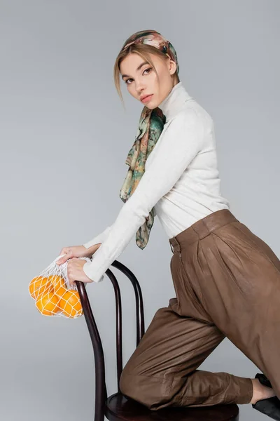 Mujer con estilo en cuello alto y pañuelo posando con silla y bolsa de red con naranjas aisladas en gris - foto de stock