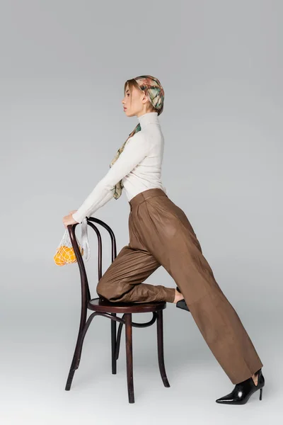 Vista lateral de mujer en pantalones posando con silla y bolsa de malla con naranjas sobre fondo gris - foto de stock