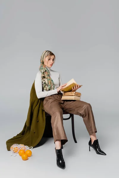 Pleine longueur de femme assise avec des livres vintage près du manteau vert et des oranges sur fond gris — Photo de stock