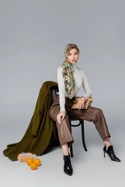 Trendy woman with vintage books sitting on chair near green coat and net bag with oranges on grey background — Stock Photo