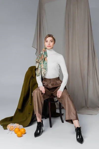 Mujer elegante sentada en silla cerca de abrigo verde y naranjas frescas sobre fondo gris con cortinas - foto de stock