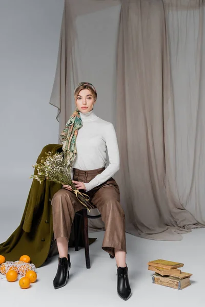 Femme dans des vêtements élégants assis avec des fleurs près des livres vintage et des oranges sur fond gris avec draperie — Photo de stock