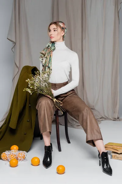 Молодая и стильная женщина, сидящая с букетом рядом с зеленым пальто и апельсинами на сером фоне — стоковое фото
