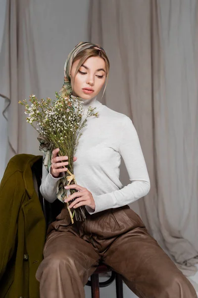 Elegante donna in dolcevita bianco seduta con bouquet di fiori di gypsophila su sfondo grigio — Foto stock