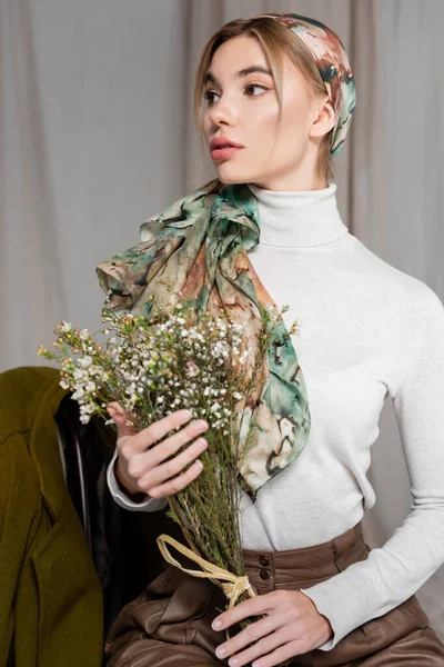 Mujer joven en cuello alto blanco sosteniendo flores de gypsophila y mirando hacia otro lado sobre fondo gris - foto de stock