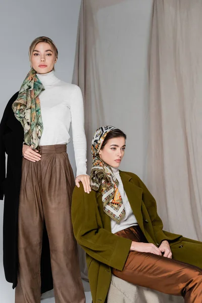 Woman in kerchief looking at camera near elegant friend sitting on grey background with drapery — Stock Photo