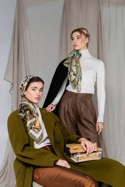 Woman in patterned kerchief sitting with vintage books near friend on grey draped background — Stock Photo