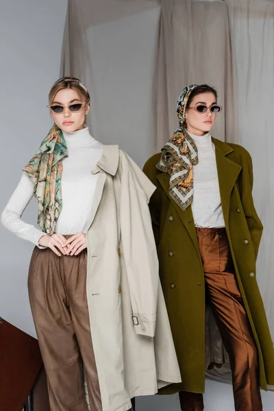 Women in stylish kerchiefs and sunglasses posing on grey background with cloth — Stock Photo