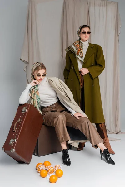 Trendy woman sitting on vintage suitcase near friend and oranges on floor on grey draped background — Stock Photo