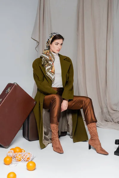 Élégante femme assise sur une valise vintage près d'oranges et de livres vintage sur fond drapé gris — Photo de stock