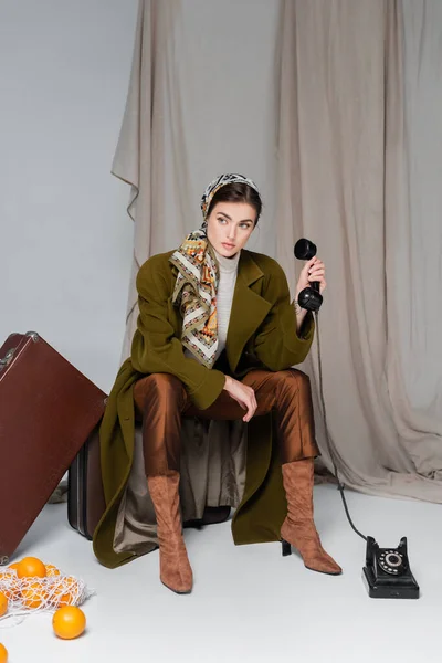 Elegante donna con telefono cellulare retrò seduto sulla valigia vicino arance su sfondo grigio con stoffa — Foto stock