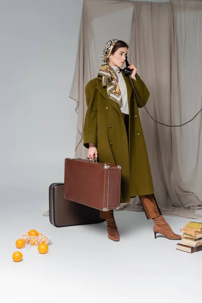 Femme à la mode avec valise vintage appelant au téléphone près des livres et des oranges sur fond drapé gris — Photo de stock