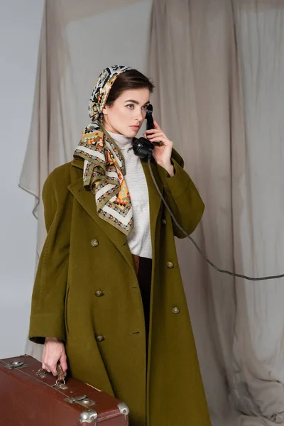 Woman in green coat and patterned kerchief holding suitcase and talking on vintage phone on grey draped background — Stock Photo