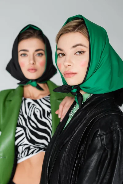 Stylish woman in green kerchief looking at camera near blurred friend isolated on grey — Stock Photo