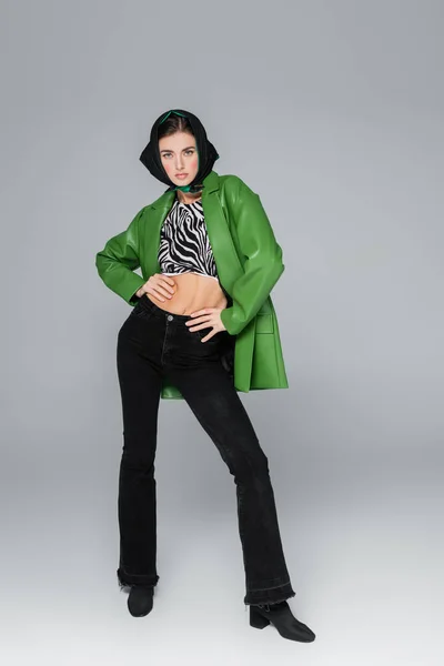 Полная длина модели в зеленой куртке и черных джинсах, держащих руки на талии на сером фоне — стоковое фото