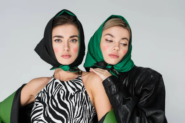 Woman in animal print top and black kerchief looking at camera near friend isolated on grey — Stock Photo