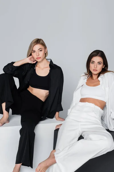 Sensuales mujeres descalzas en ropa blanca y negra posando sobre cubos aislados en gris - foto de stock