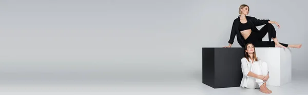 Piena lunghezza della donna seduta su cubi in bianco e nero vicino amico sul pavimento su sfondo grigio, banner — Foto stock