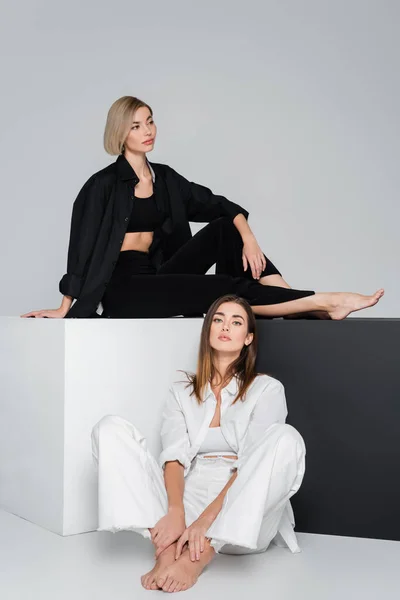 Barefoot woman in white clothes sitting on floor near friend on cubes on grey background — Stock Photo