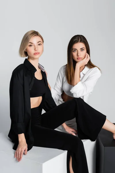 Mujeres en ropa blanca y negra sentadas en cubos y mirando a la cámara aislada en gris - foto de stock