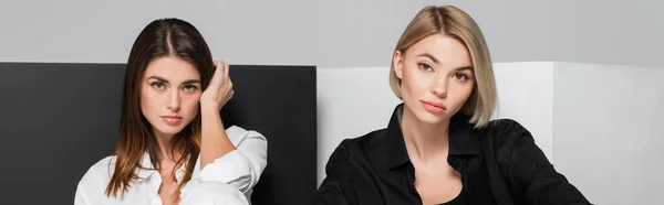 Brunette and blonde women looking at camera near black and white cubes isolated on grey, banner — Stock Photo