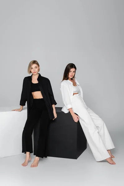 Longitud completa de las mujeres descalzas en ropa elegante posando cerca de cubos negros y blancos sobre fondo gris - foto de stock