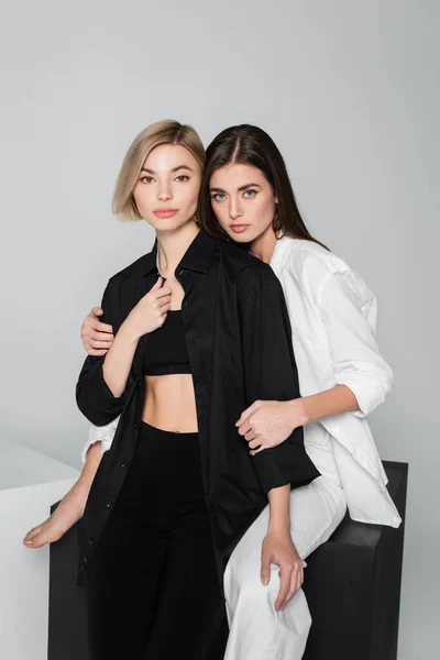 Brunette woman hugging friend in black clothes while sitting on cube isolated on grey — Stock Photo