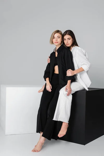 Brunette woman hugging friend wearing black clothes while sitting on cube on grey background — Stock Photo