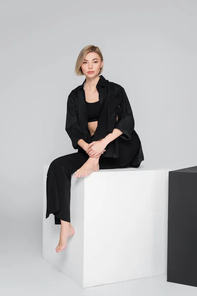 Tutta la lunghezza della donna scalza in abiti neri seduta sul cubo su sfondo grigio — Foto stock